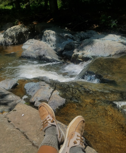 A pair of feet in sneakers resting on a rock by a flowing stream in a natural setting.