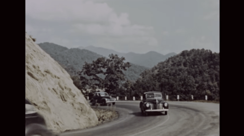 A vintage car navigates a winding mountain road with lush hills in the background under a partly cloudy sky.