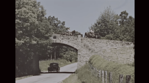 A vintage car drives under a stone bridge with horses standing on top, surrounded by lush greenery.