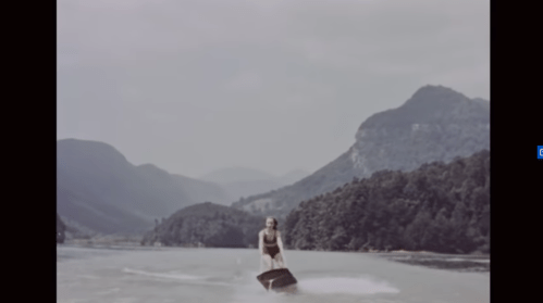A person water skiing on a lake surrounded by mountains and trees on a sunny day.