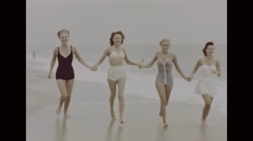 Four women in vintage swimsuits joyfully walk hand-in-hand along a beach, with gentle waves in the background.