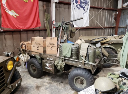 A military vehicle with supplies, including boxes labeled "Meal Combat Individual," in a hangar with flags in the background.