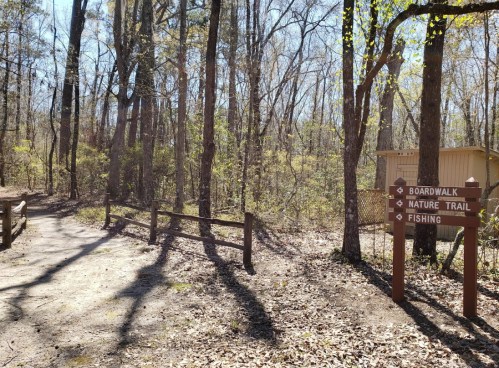 A wooded area with a signpost indicating directions to a boardwalk, nature trail, and fishing. Sunlight filters through trees.