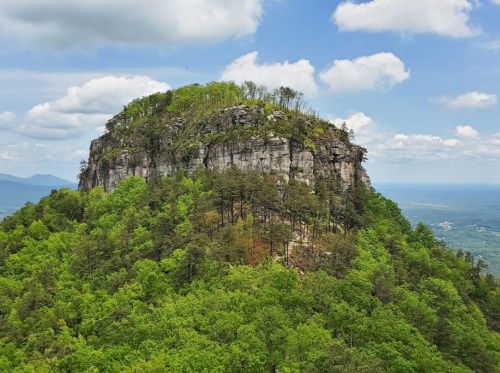 A rocky mountain peak surrounded by lush green trees under a partly cloudy sky.