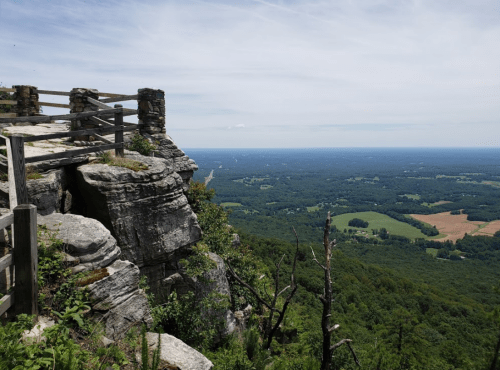 A scenic view from a rocky overlook, showcasing lush green valleys and distant hills under a clear blue sky.