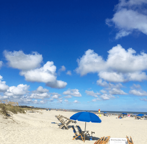 A sunny beach scene with blue skies, fluffy clouds, and an umbrella over lounge chairs on the sandy shore.