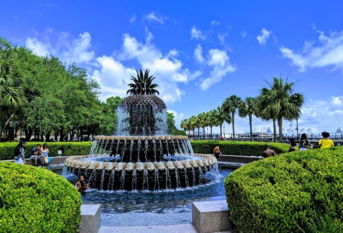 A tiered fountain surrounded by lush greenery and palm trees, with people enjoying the park on a sunny day.