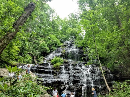 A scenic waterfall surrounded by lush green trees, with people exploring the area at its base.