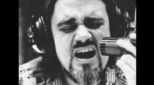 A man with long hair and a beard passionately sings into a microphone, wearing headphones in a black and white setting.