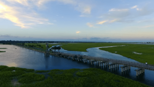 Aerial view of a wooden pier extending over a calm river, surrounded by lush green marshland and a colorful sky.