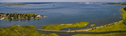 Aerial view of a coastal landscape with a winding pier, green marshlands, and boats on the water under a clear sky.