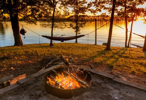 A serene lakeside scene at sunset, featuring a campfire, hammock, and trees reflecting on the water.