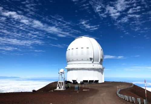 A large white observatory dome on a mountain peak, surrounded by clouds and a blue sky.