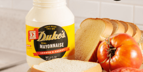 A jar of Duke's mayonnaise next to slices of bread, a tomato, and an apple on a kitchen countertop.