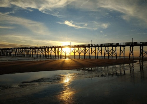 Sunset over a pier, casting shadows on the beach with calm waters reflecting the sky.