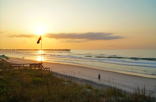 Sunset over a beach with gentle waves, a pier in the distance, and a person walking along the shore.