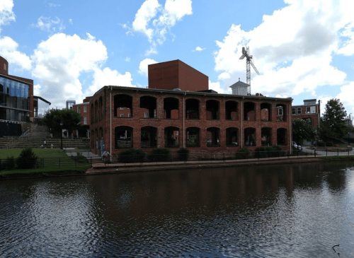 Historic brick building with arched windows by a calm river, under a blue sky with scattered clouds.