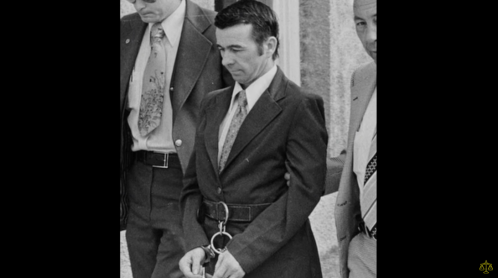 A man in a suit is handcuffed and flanked by two individuals in formal attire, appearing somber.