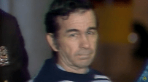 A man with dark hair and a striped sweater looks solemn, surrounded by law enforcement in a blurred background.