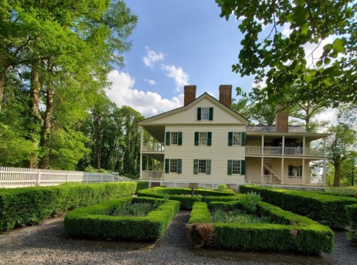 A historic two-story house with green shutters, surrounded by manicured gardens and trees under a blue sky.