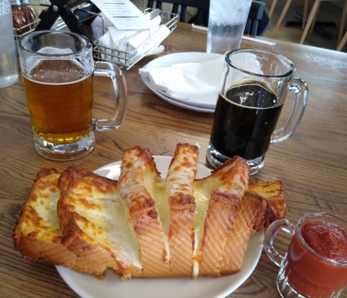 Two glasses of beer beside a plate of cheesy bread with a small cup of marinara sauce.