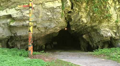 Entrance to a rocky cave with a colorful totem pole beside it, surrounded by greenery.