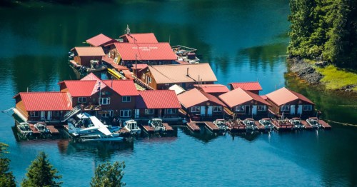 Aerial view of colorful floating cabins on a calm lake, surrounded by lush greenery and boats docked nearby.