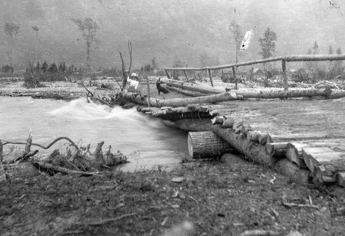 A rustic wooden bridge over a rushing river, surrounded by trees and fog, depicting a natural landscape.