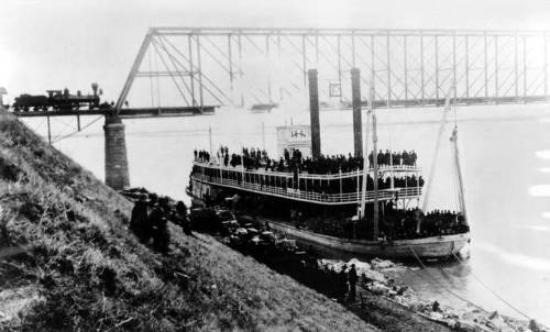 Historic black-and-white photo of a steamboat with passengers near a bridge, with a train passing overhead.