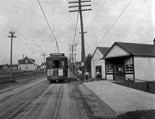 A vintage streetcar on tracks near a grocery store, with power lines and houses in the background.