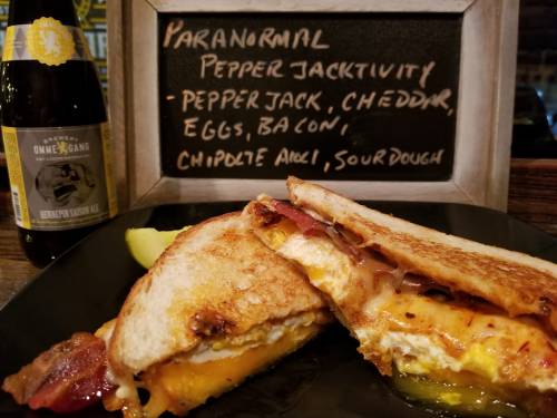 A grilled sandwich with pepper jack cheese, eggs, bacon, and chipotle aioli, served with a pickle and a beer.