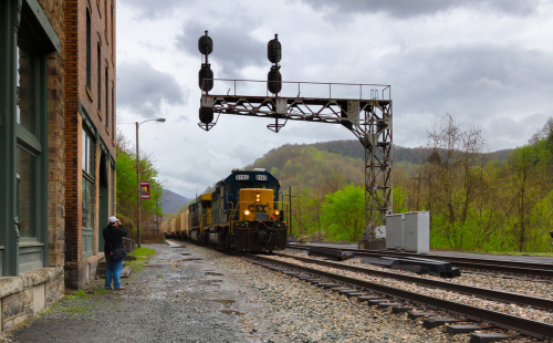 A CSX train passes under a signal bridge near a brick building, with green hills in the background and a cloudy sky.