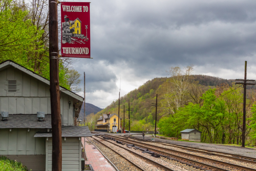 A sign reading "Welcome to Thurmond" near train tracks, with green hills and cloudy skies in the background.
