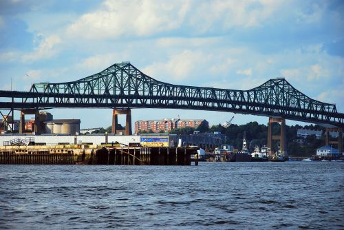 A large green bridge spans over a river, with buildings and boats visible in the background under a cloudy sky.