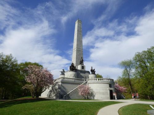 A tall stone monument with statues, surrounded by trees and blooming flowers under a blue sky with clouds.