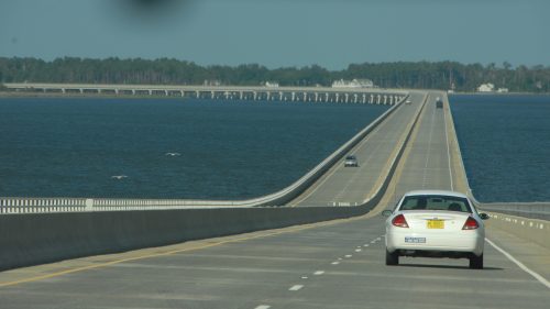 A car drives on a long bridge over water, with trees and a distant shoreline visible in the background.