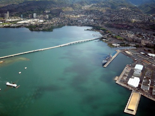 Aerial view of a harbor with a bridge, boats, and buildings along the shoreline, surrounded by green hills.