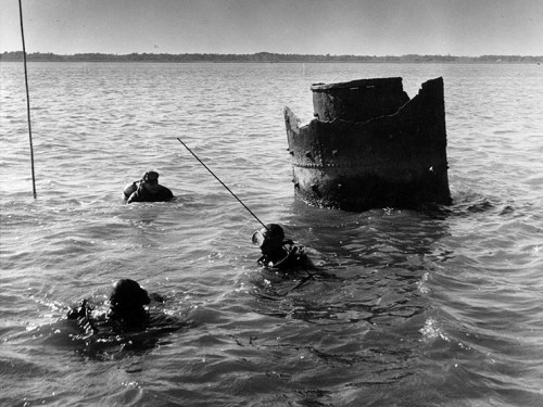 Three divers in water near a submerged structure, with a clear sky and distant shoreline in the background.