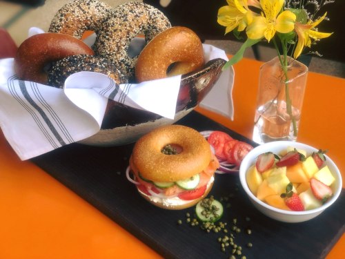 A basket of assorted bagels, a bagel sandwich with veggies, and a fruit bowl on a vibrant orange table.