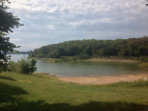 A serene lakeside view with a sandy shore, surrounded by lush green trees under a cloudy sky.