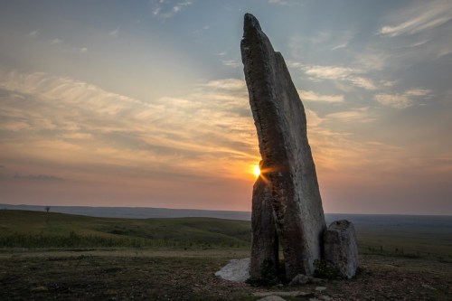 A tall stone monolith stands against a sunset, casting a shadow over a grassy landscape.