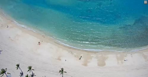 Aerial view of a sandy beach with turquoise water and a few people enjoying the sun. Palm trees line the shore.