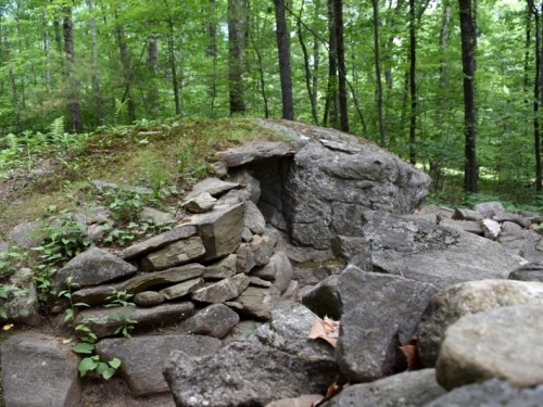 A small stone structure partially covered by moss, surrounded by trees and rocks in a forested area.