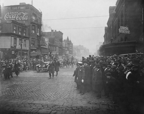 Historic black-and-white photo of a crowded street with onlookers, vintage cars, and early 20th-century buildings.