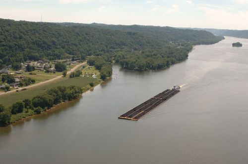 A barge navigates a wide river, surrounded by green hills and a small town along the shore.
