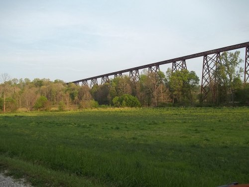 A tall, rusted railway trestle spans a green field, surrounded by trees under a clear sky.