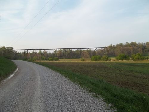 A gravel road leads to a green field with trees, under a large railway bridge in the distance.
