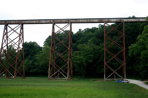 A tall, rusty railway trestle bridge stands over a grassy area, with a car parked nearby and trees in the background.