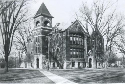 Historic brick building with a tower, surrounded by bare trees and a grassy area, captured in black and white.