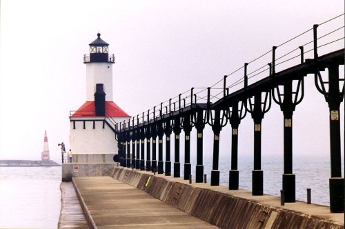 A lighthouse stands at the end of a pier, with a foggy sea in the background and another lighthouse visible in the distance.